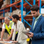 People standing in a warehouse with a man holding a tablet and two people in the background on a computer